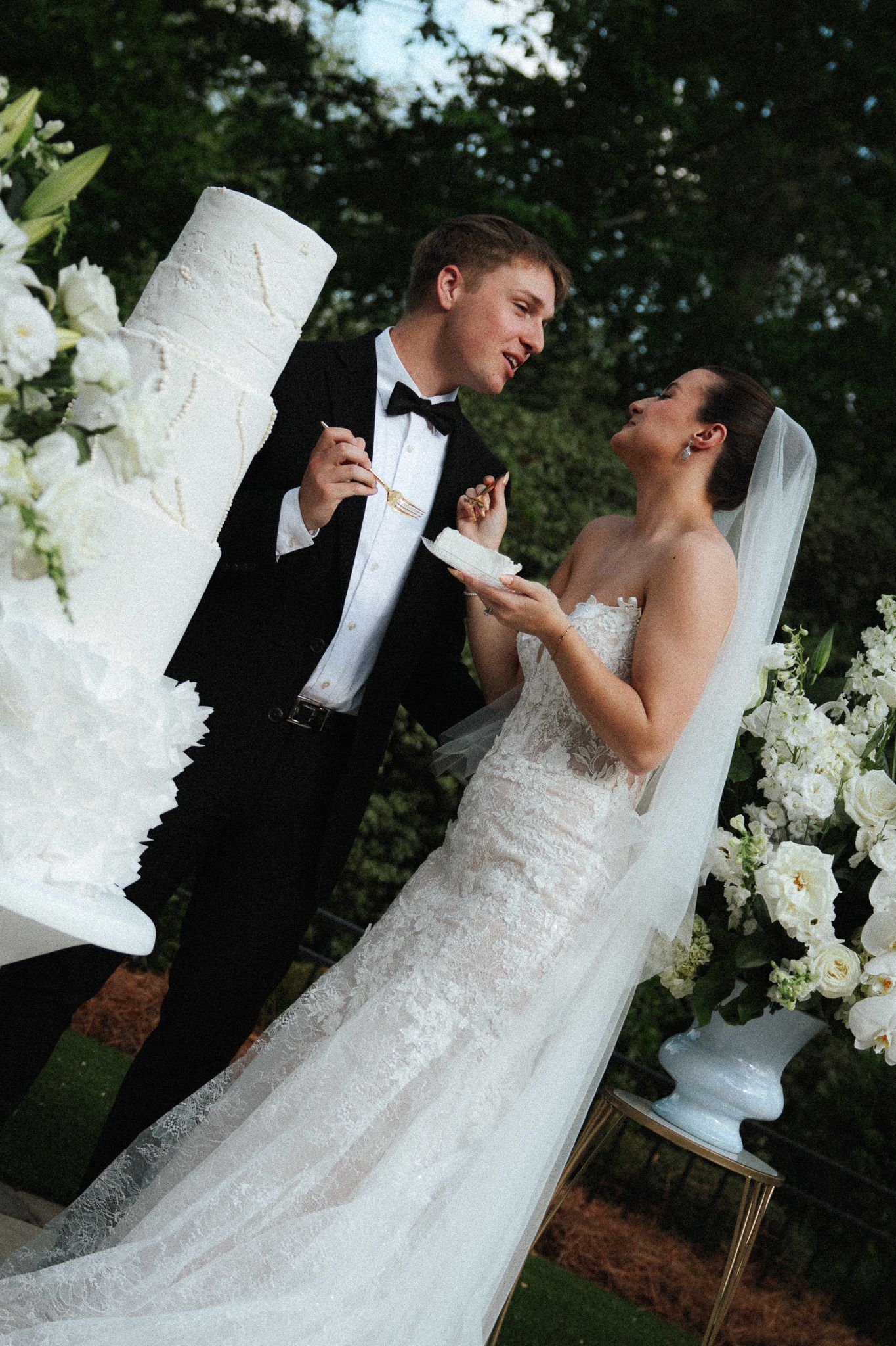 Groom feeds bride wedding cake as they laugh together beside elegant white floral decorations outdoors.