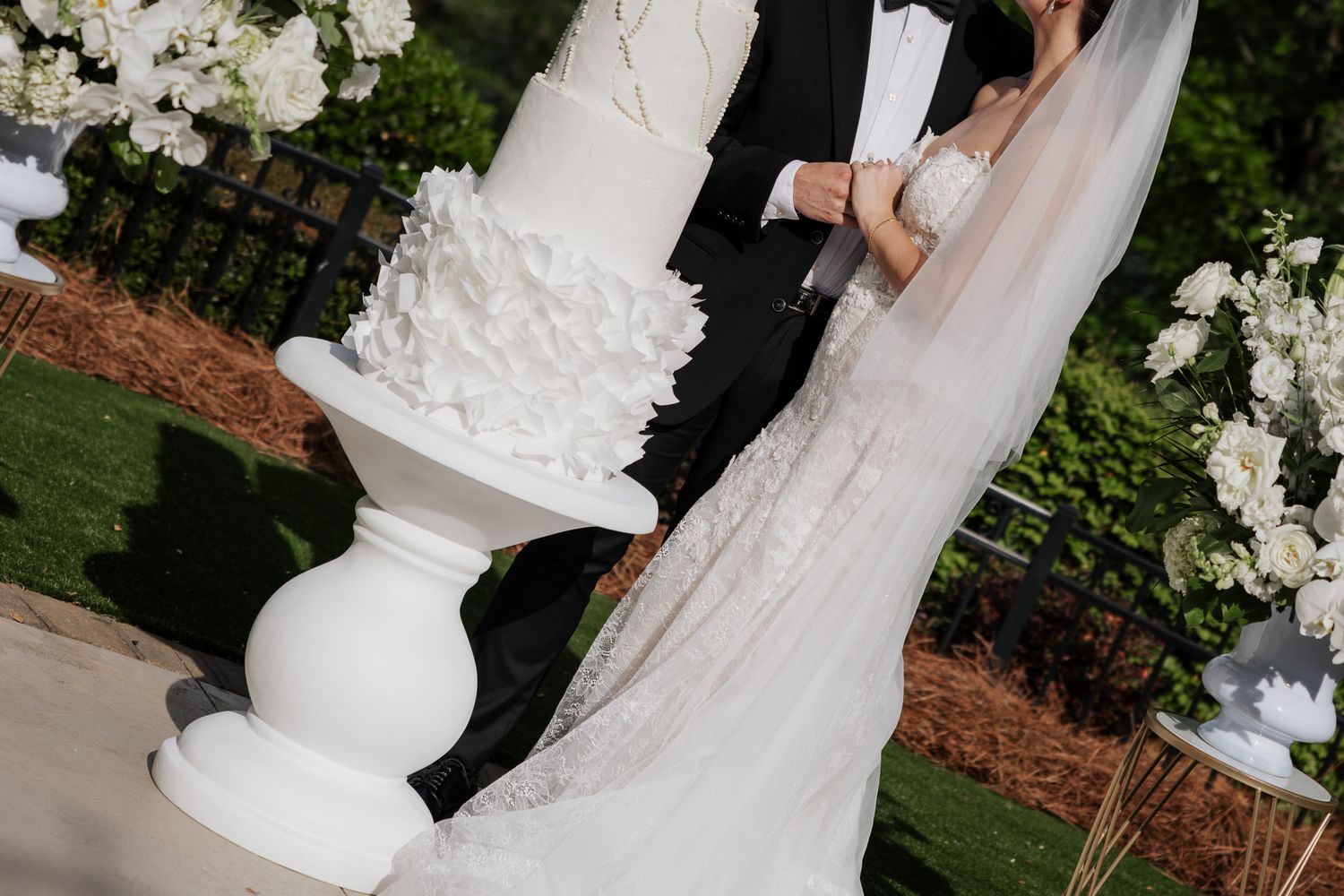 Close-up of couple cutting a stunning white ruffled tiered wedding cake on a white pedestal stand outdoors.