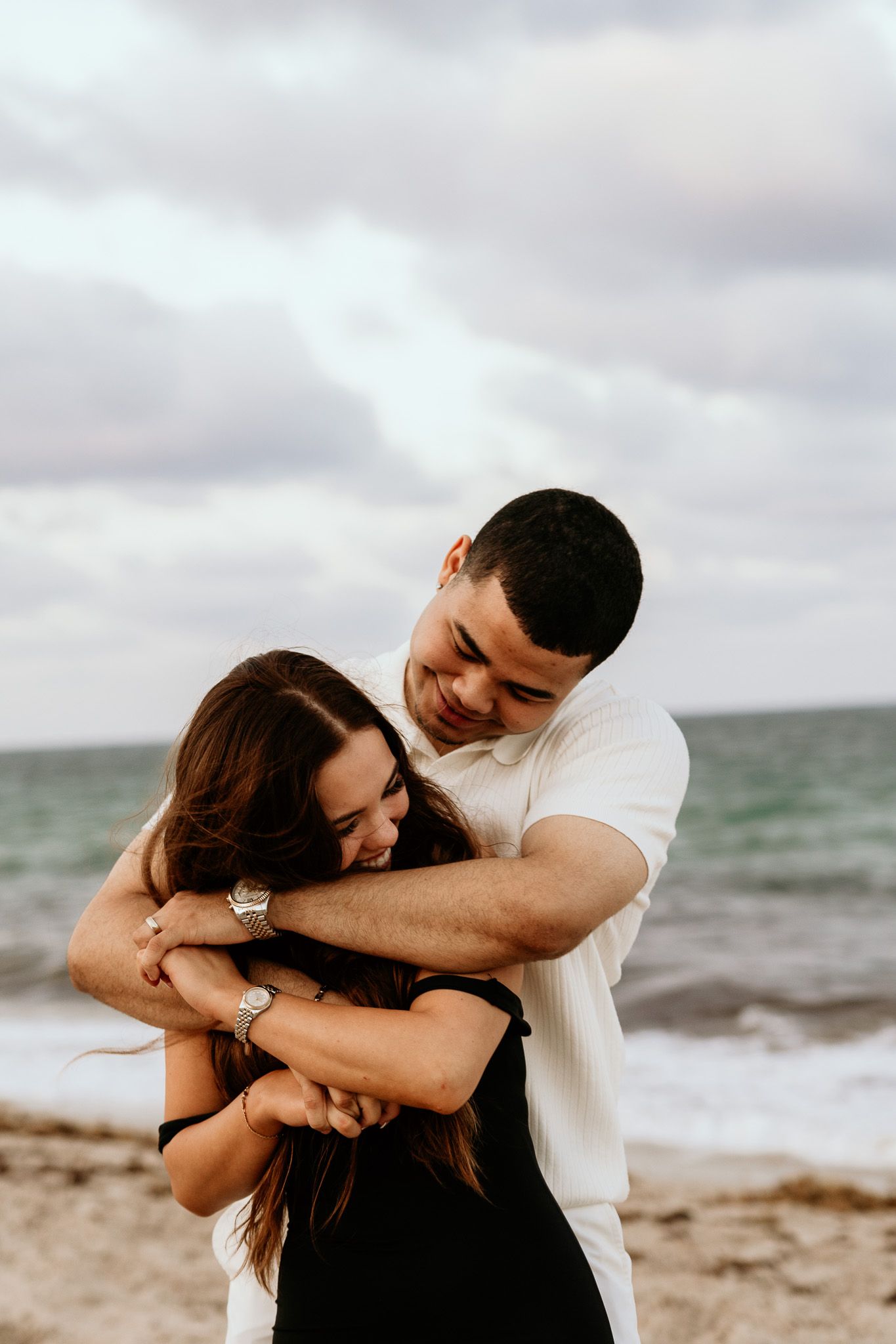Couple laughing and embracing on sandy beach with ocean waves and cloudy sky in background.