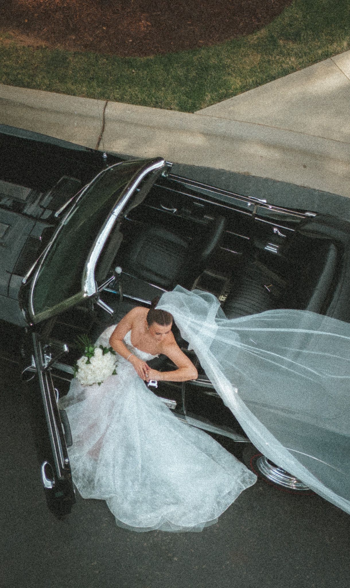 Aerial view of bride in lace gown with flowing veil beside a classic black convertible car.