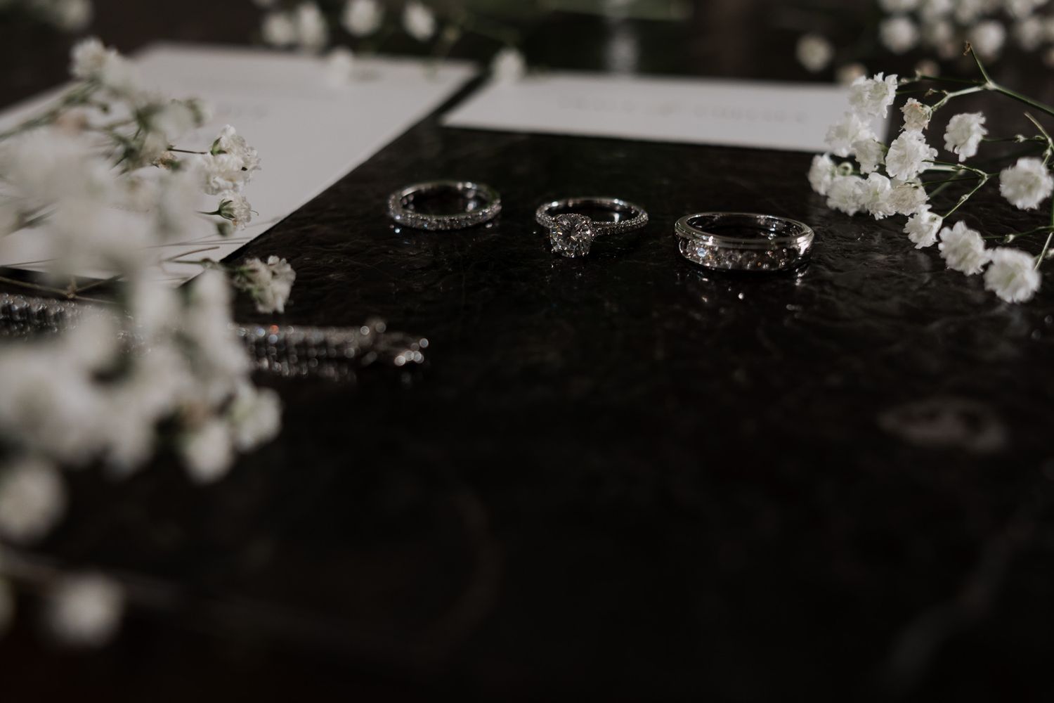 Black and white moody flat lay of wedding rings, bracelet, and baby's breath flowers on dark surface.