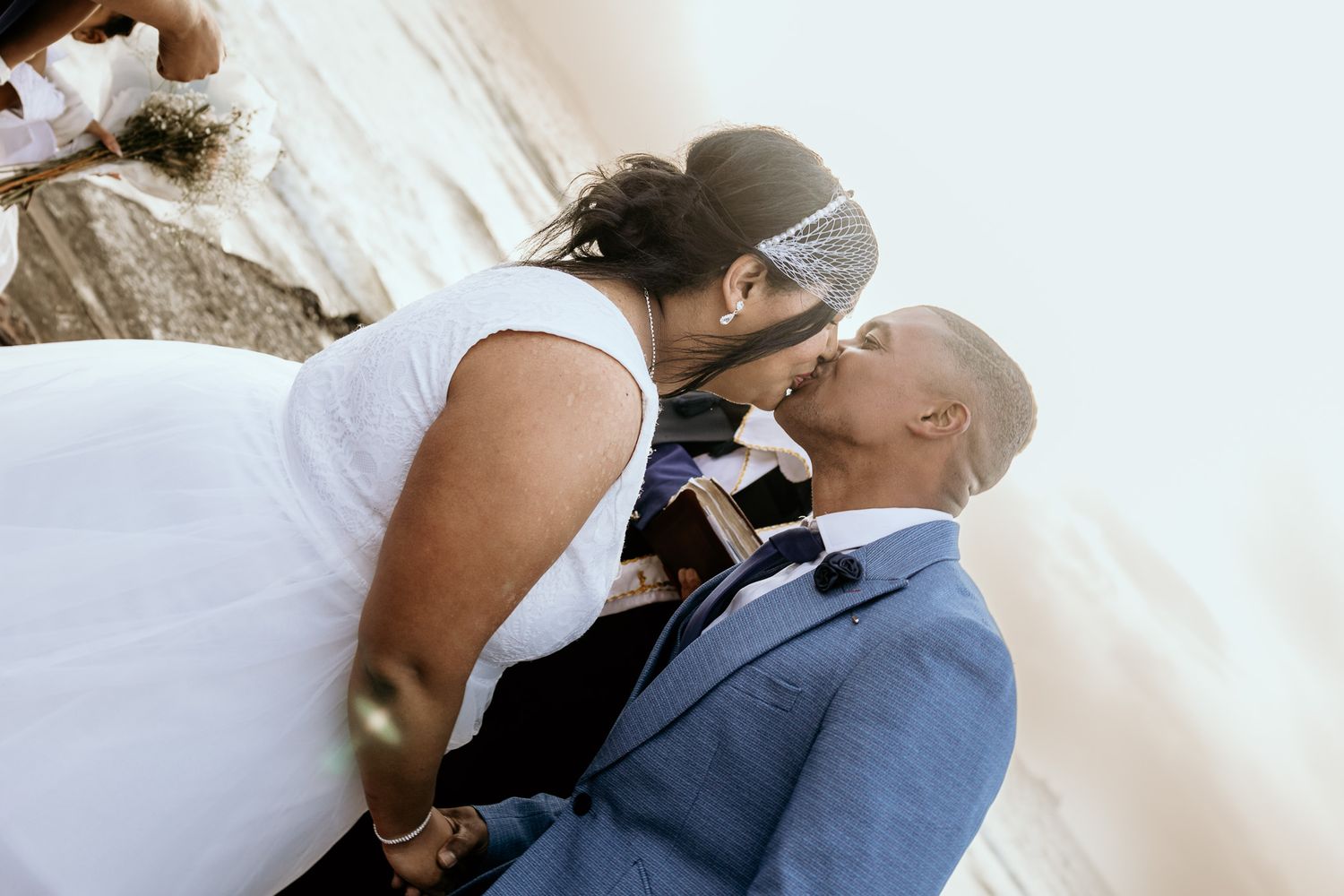 Bride in birdcage veil sharing a tender kiss with groom in blue suit during beach wedding ceremony.