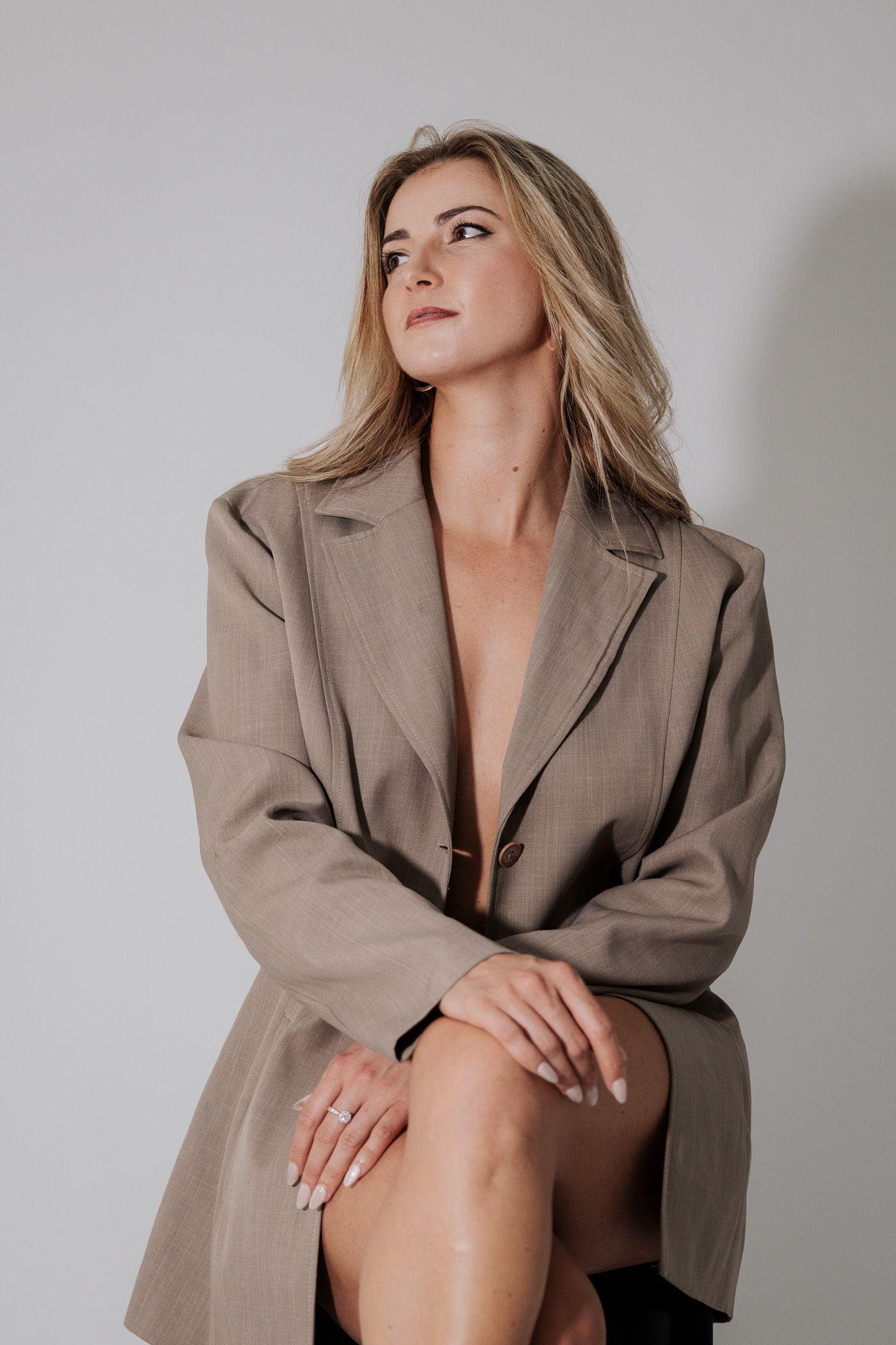 Blonde woman posing in oversized taupe blazer seated against white studio background.
