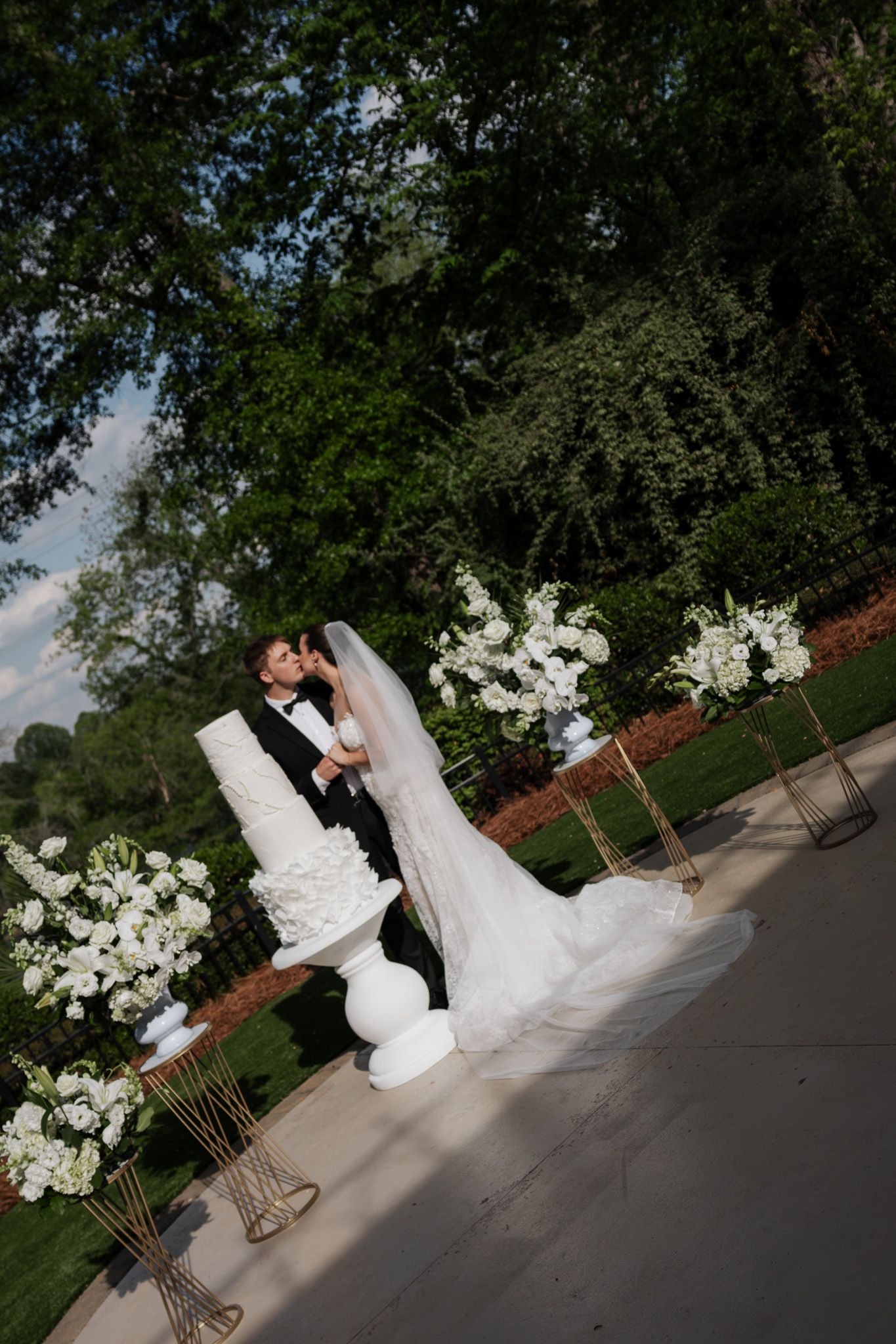 Bride and groom share a romantic kiss beside their elegant white wedding cake surrounded by tall white floral displays.