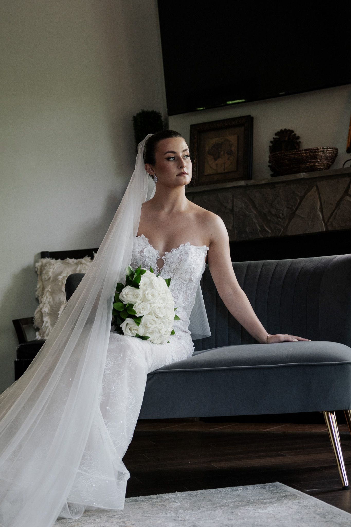 Beautiful bride in strapless lace wedding gown with long veil sits on a gray sofa holding white rose bouquet.