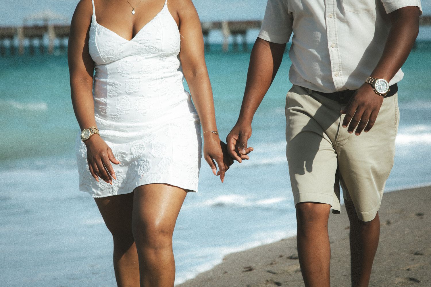 Close-up of a couple holding hands while walking on a sandy beach, both dressed in white with turquoise ocean behind them.