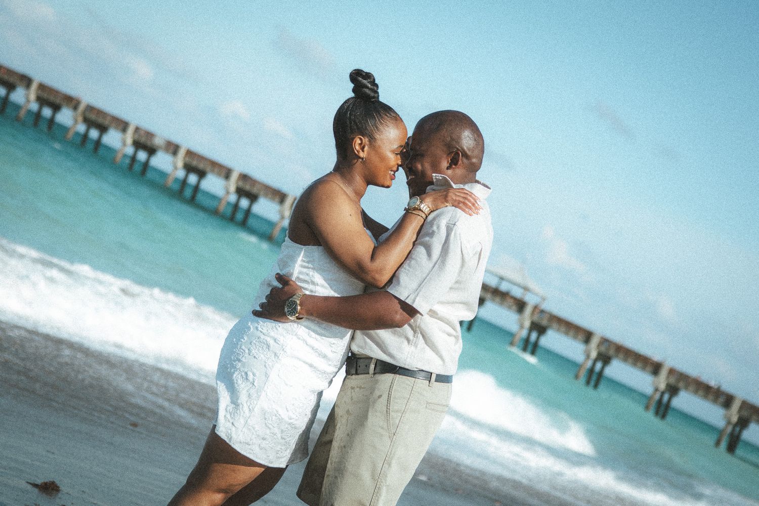A loving couple in white embracing closely on a tropical beach with clear blue water and a pier behind them.