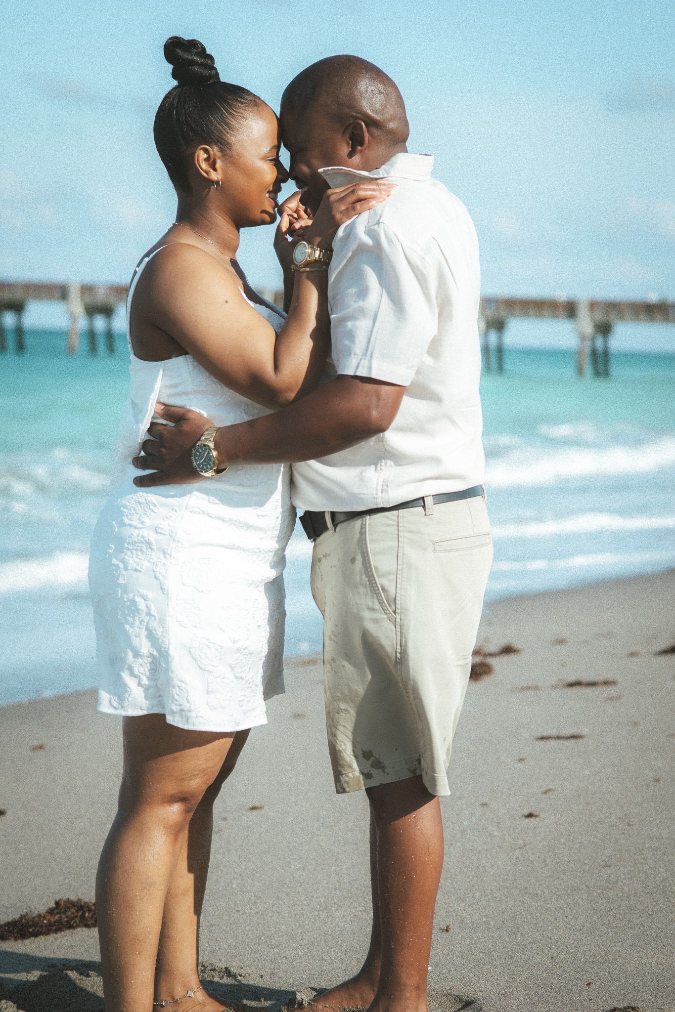 An affectionate couple dressed in white sharing an intimate moment on a sunny beach with a pier in the background.