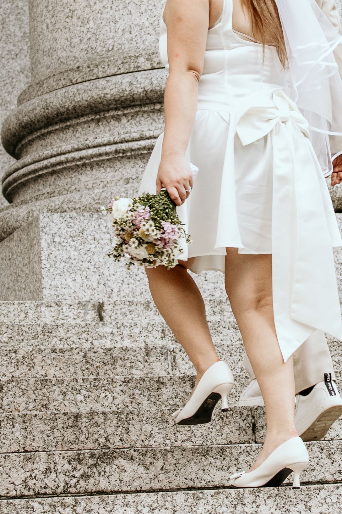 Bride in short white wedding dress and heels descending stone steps while holding a delicate floral bouquet.