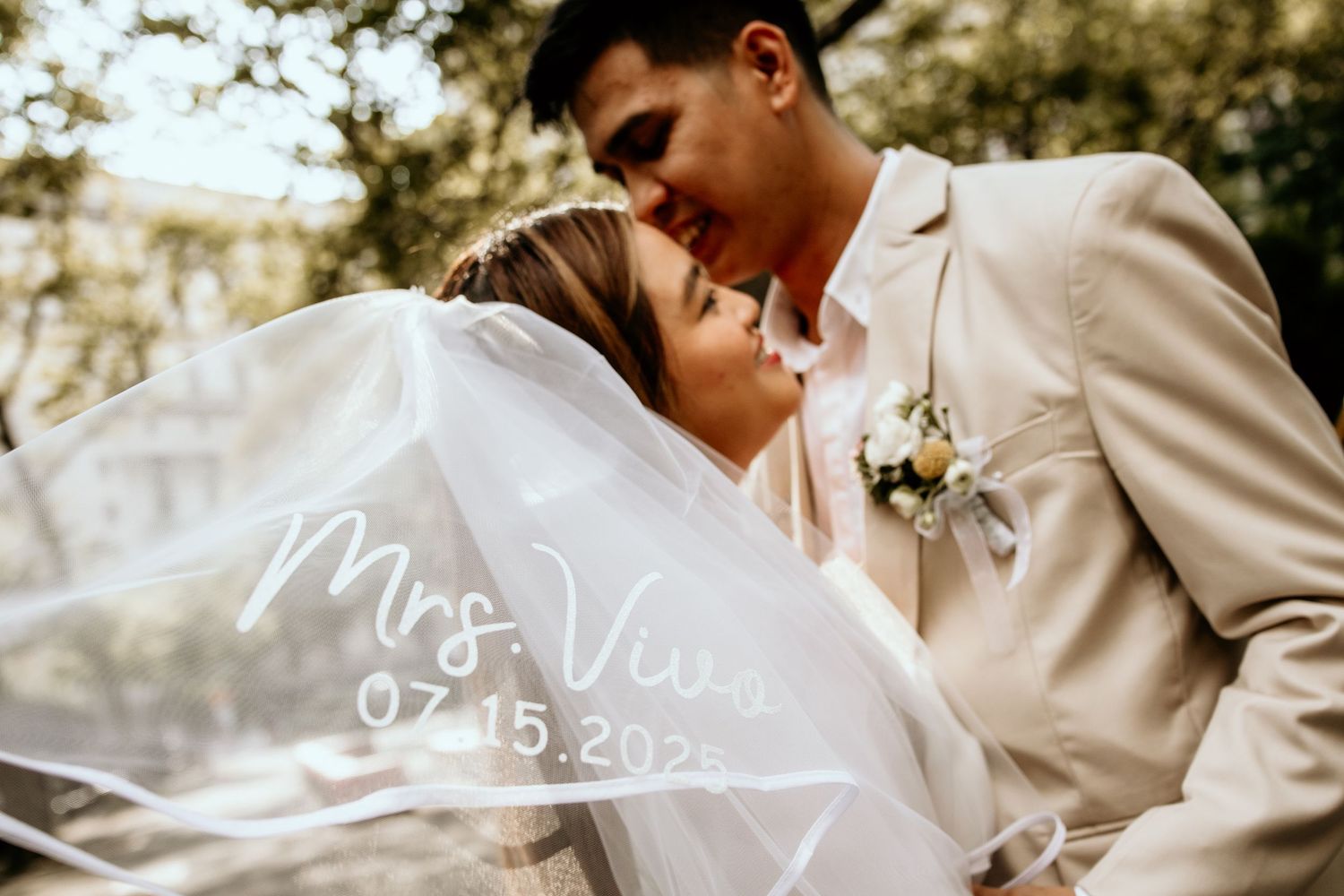 Bride and groom share a tender kiss behind veil reading Mrs. Vuu 07.15.2023 on their wedding day.