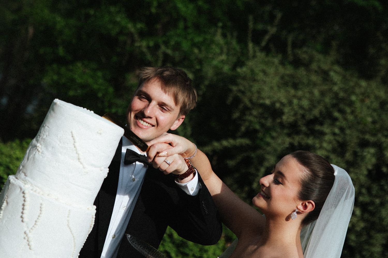 Bride and groom smiling while cutting their white wedding cake outdoors surrounded by lush green trees.