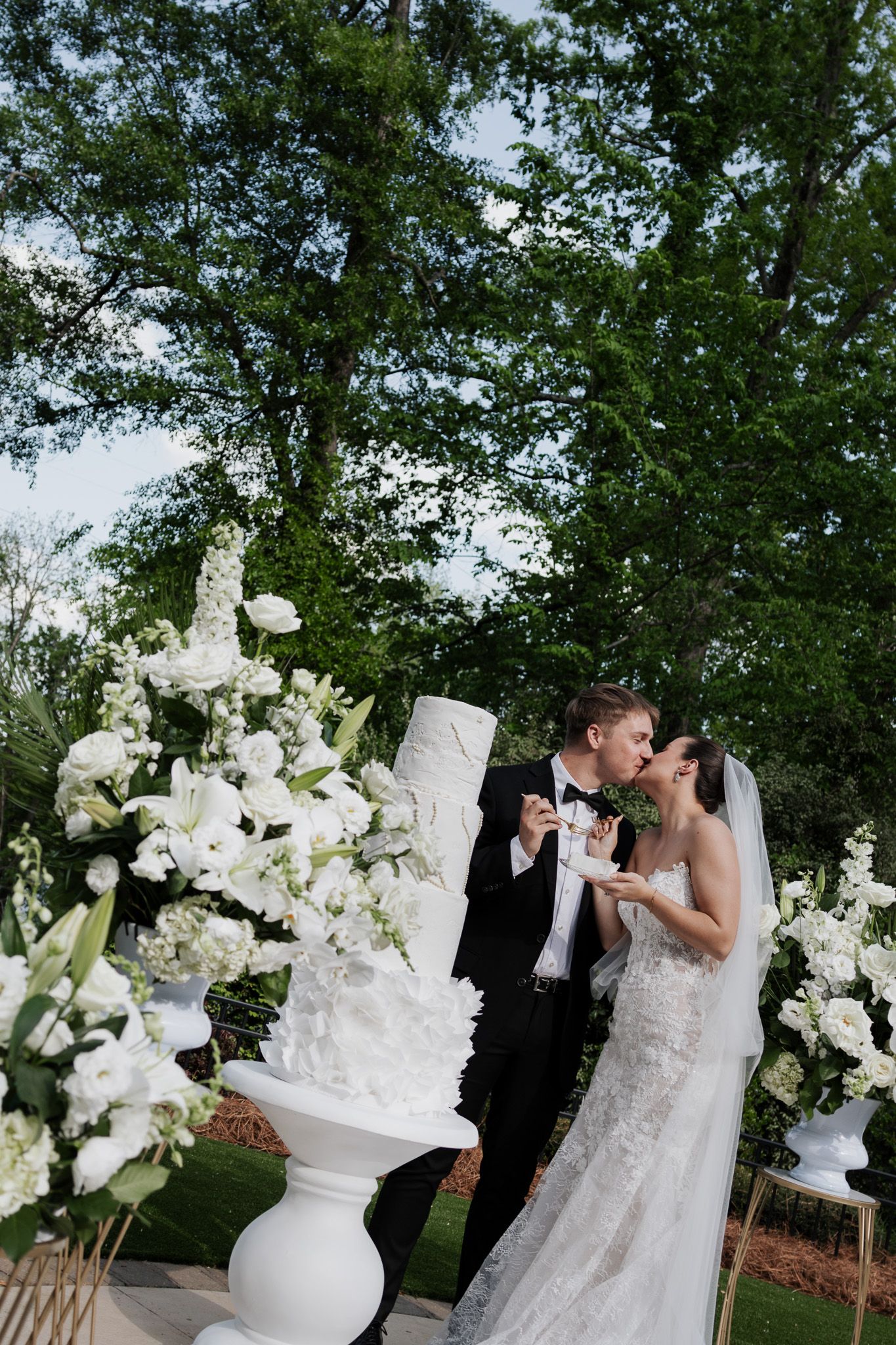Bride and groom sharing a romantic kiss beside their elegant white tiered wedding cake surrounded by white flowers.