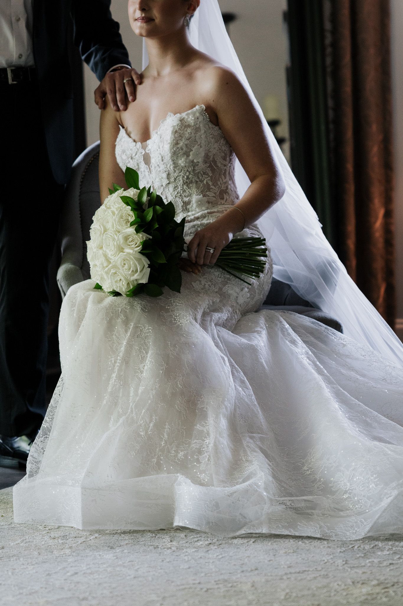 Bride in lace strapless gown holding white rose bouquet while groom stands behind her adjusting her dress.