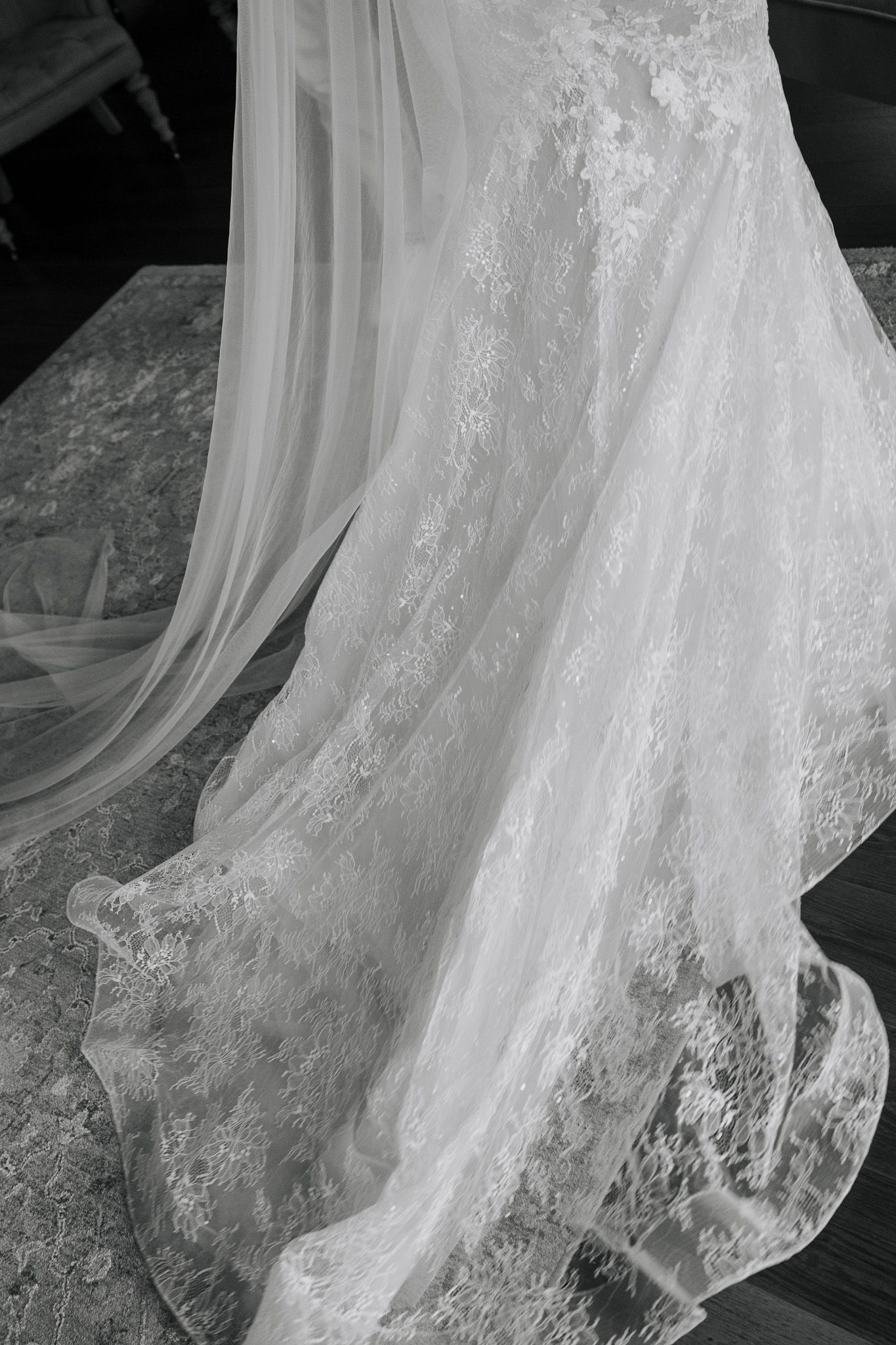 Close-up black and white detail of intricate lace and tulle wedding dress train spread across the floor.