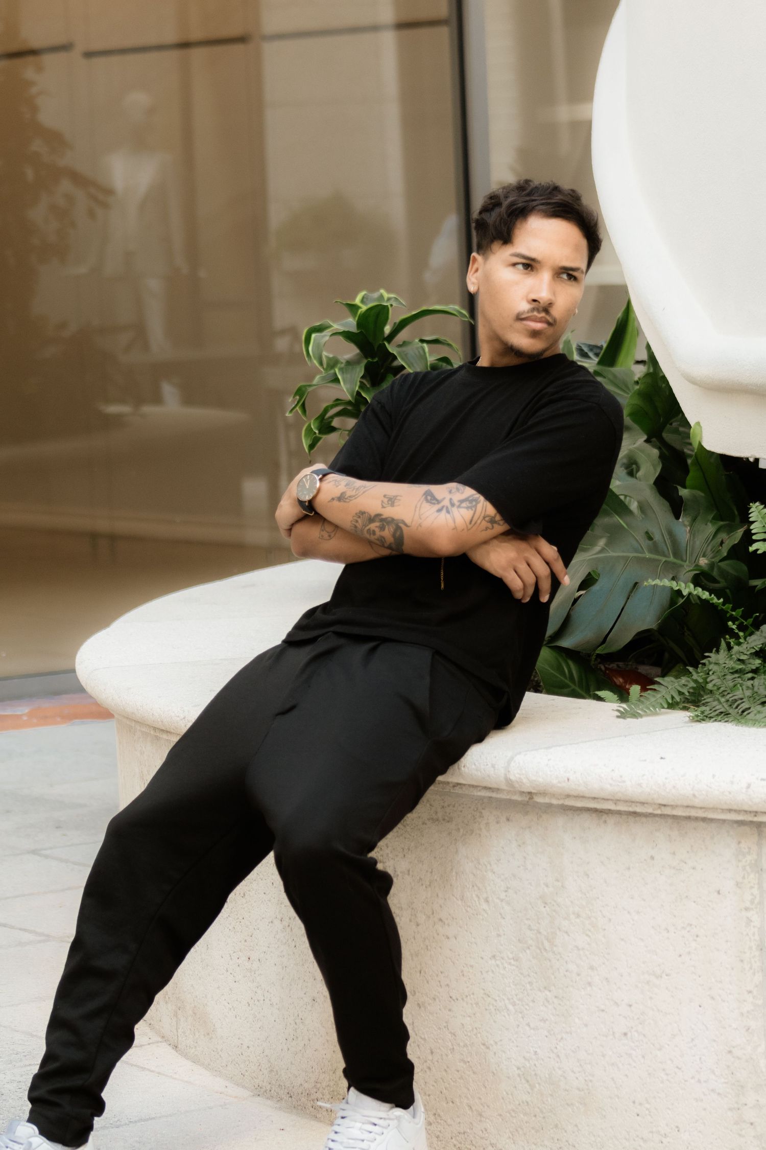 Tattooed man in all-black outfit sitting relaxed against a stone planter with tropical plants.