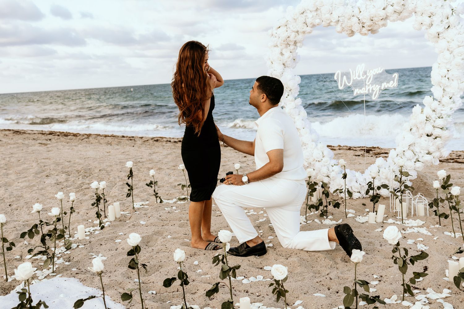 Man proposing on one knee to woman at a romantic beach setup with white roses and heart arch.