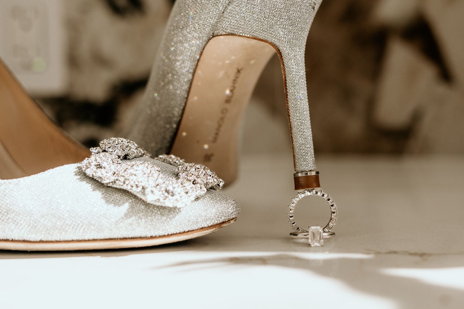 Sparkling silver bridal heels with crystal bow detail and diamond engagement ring on white floor.