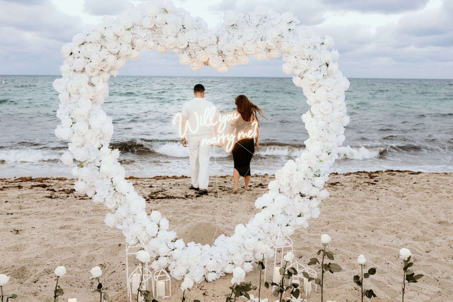 Couple walking hand-in-hand viewed through a white floral heart arch on a sandy beach.