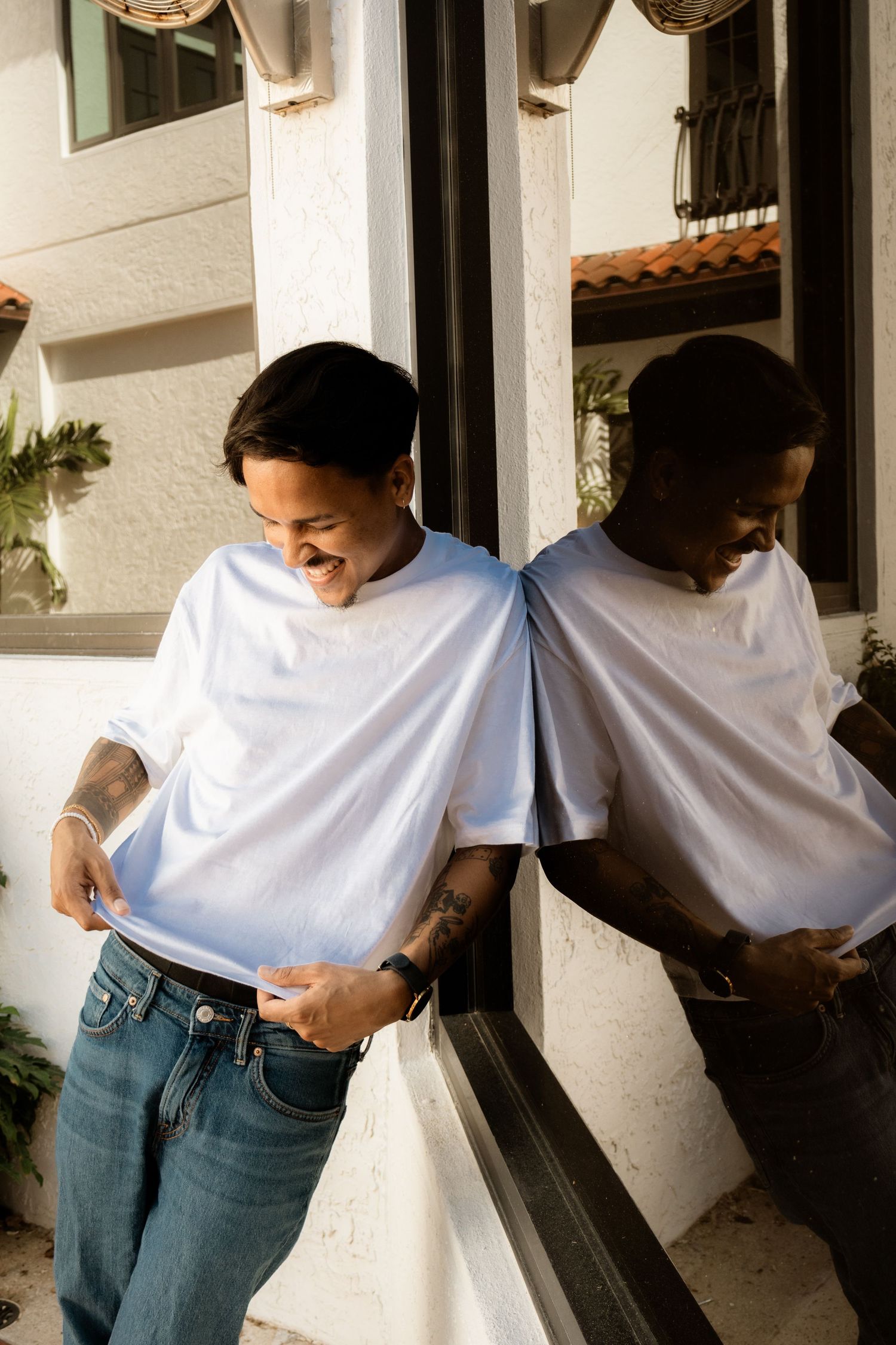 Smiling tattooed man in white tee and jeans leaning against a mirrored window outside.