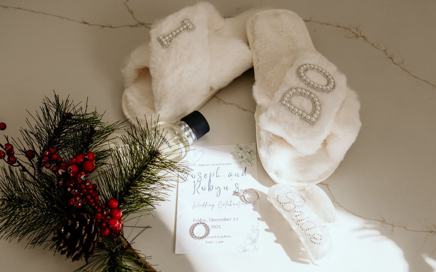 Wedding day getting ready flat lay with fluffy robe, perfume, invitation, and holiday greenery.