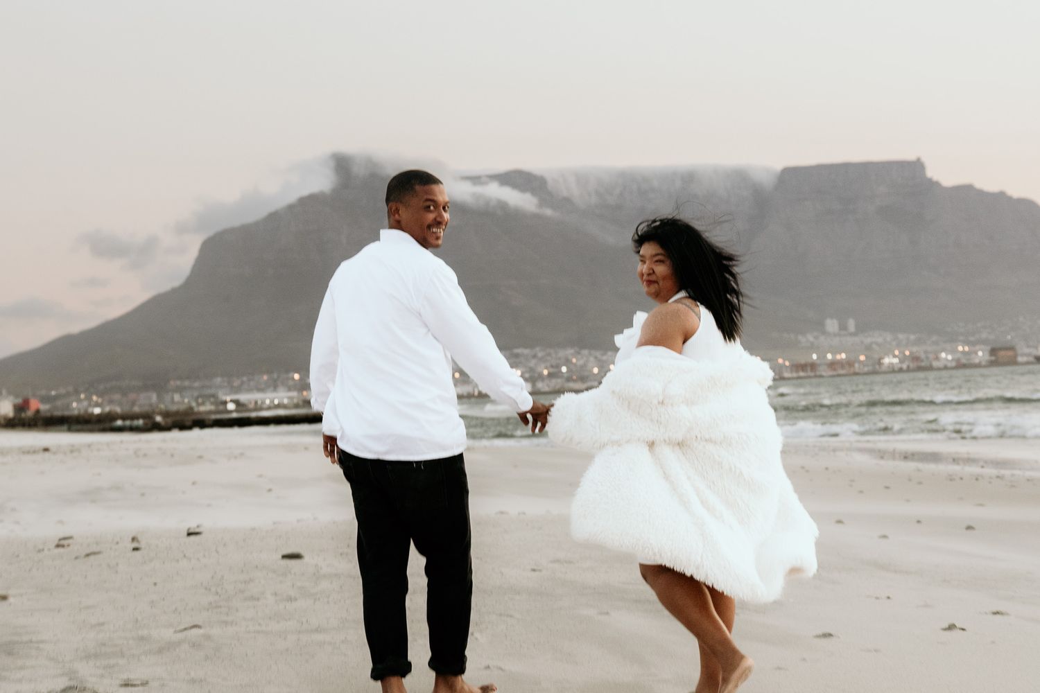 Couple in white holding hands on beach, woman in fur coat, Table Mountain behind them.