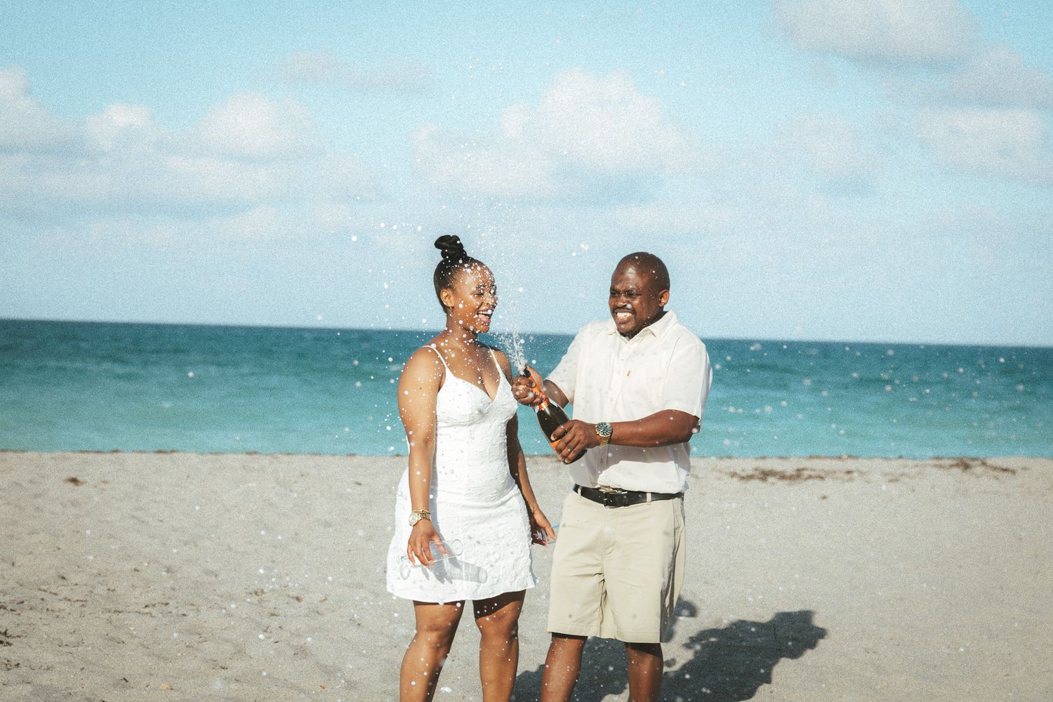 A cheerful couple in white outfits celebrating on a sunny beach by popping a bottle of champagne together.