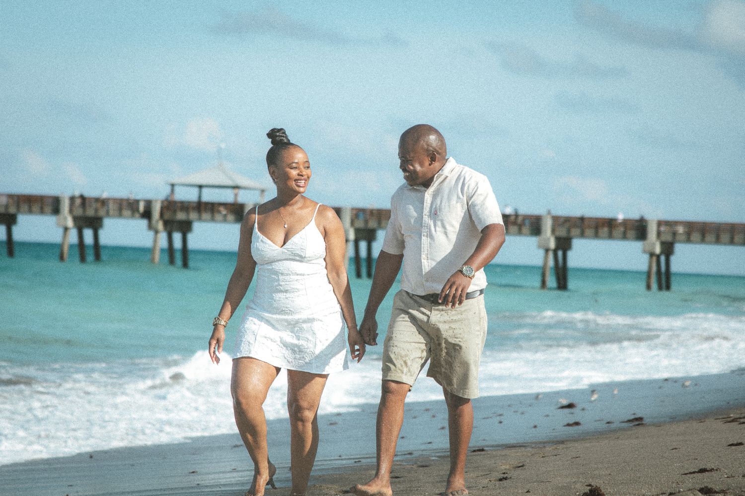 Romantic couple in white outfits stroll hand in hand along a sunny beach with turquoise waves and a distant pier.