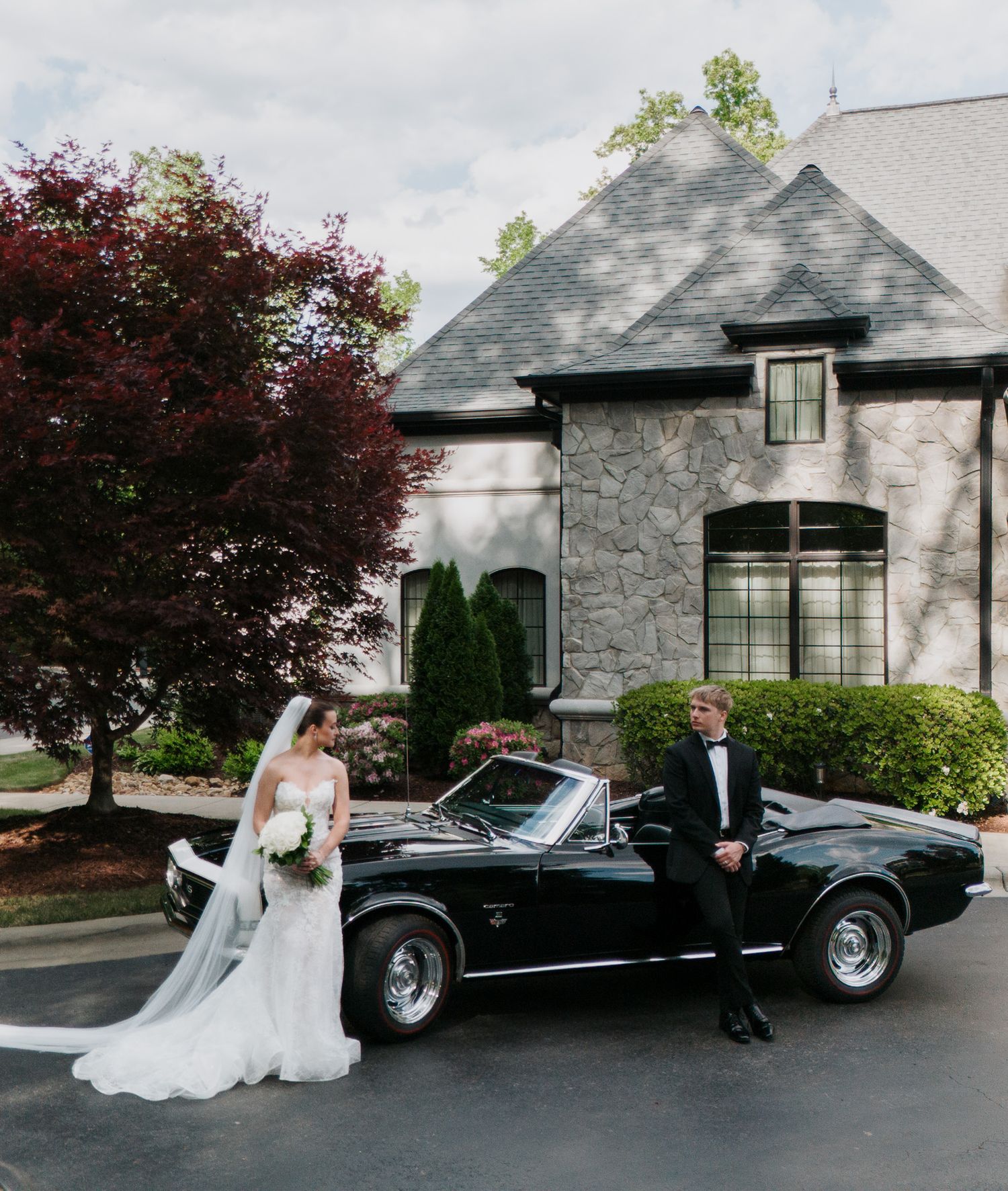 Bride and groom posing with a classic black convertible in front of an elegant stone mansion.