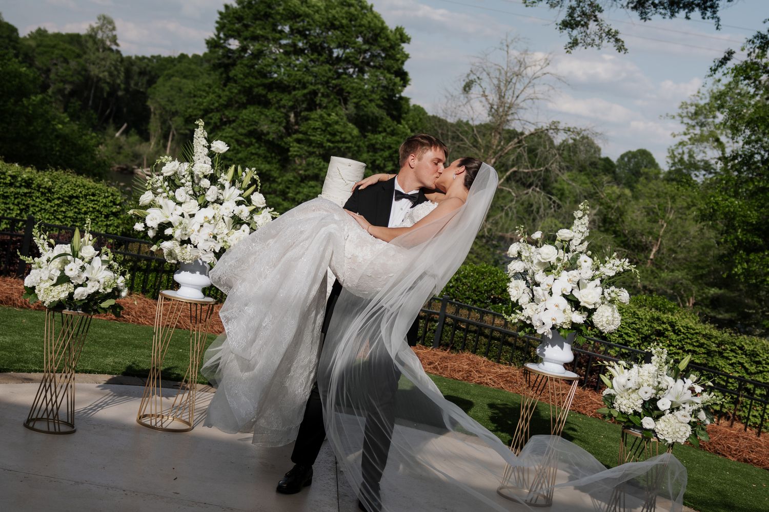 Groom dips bride in elegant white wedding gown and veil, kissing outdoors with white floral arrangements.
