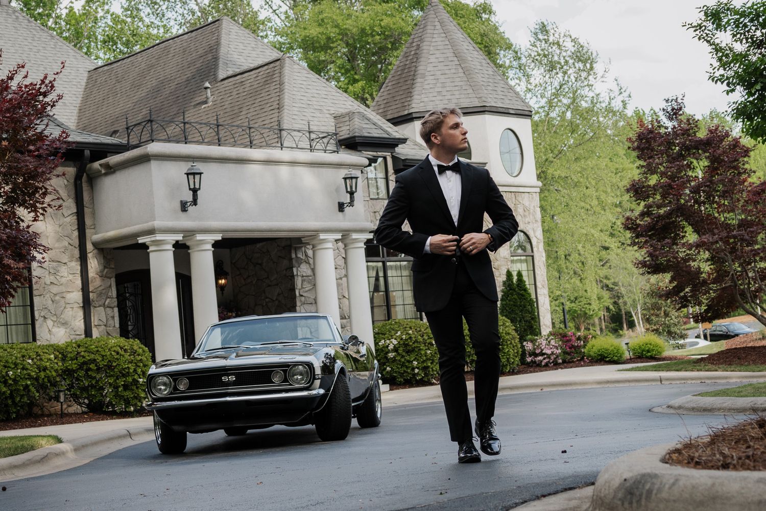 Groom in a black tuxedo walks confidently down a driveway beside a classic car in front of an elegant venue.