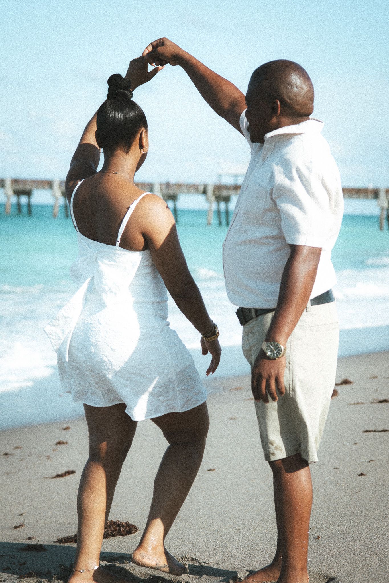 Couple dances on a sunny beach, man spinning woman in a white dress with turquoise ocean waves and a pier in background.
