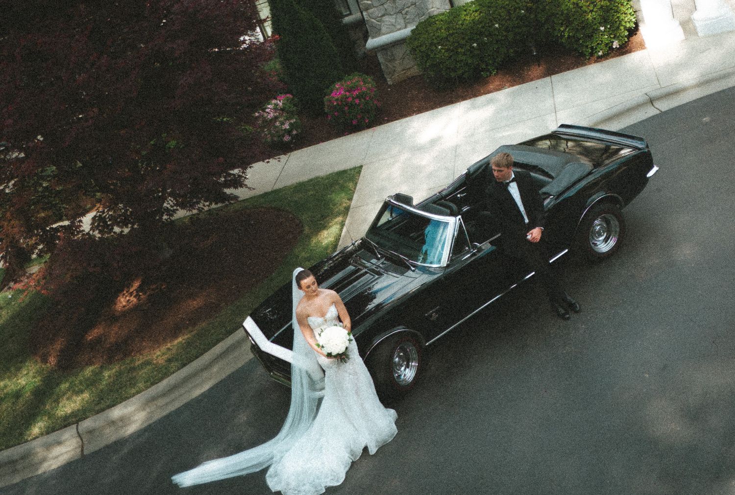 Bride and groom posing beside a classic black convertible muscle car on their wedding day.