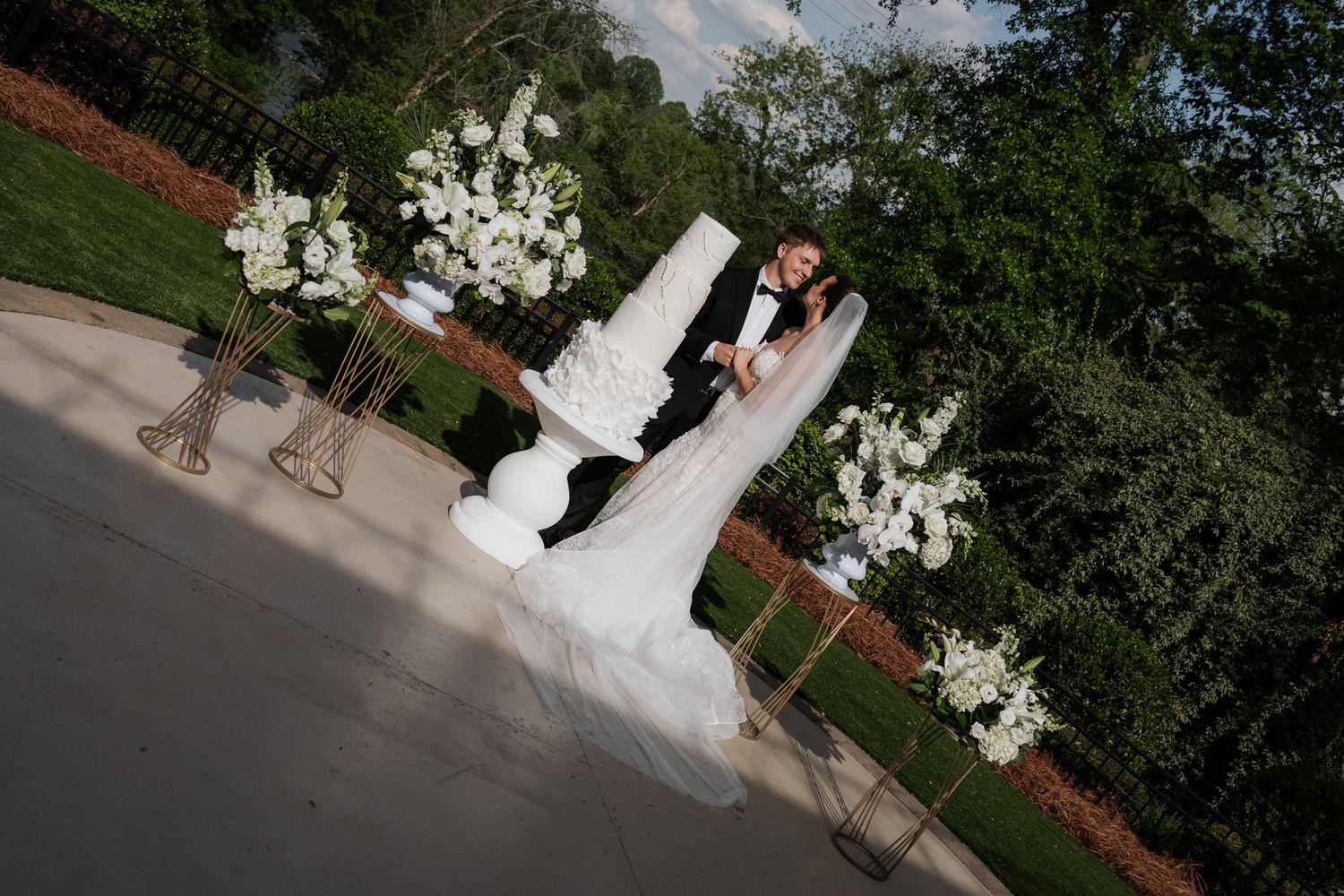 Aerial view of bride and groom kissing beside towering white wedding cake flanked by lush white floral arrangements.