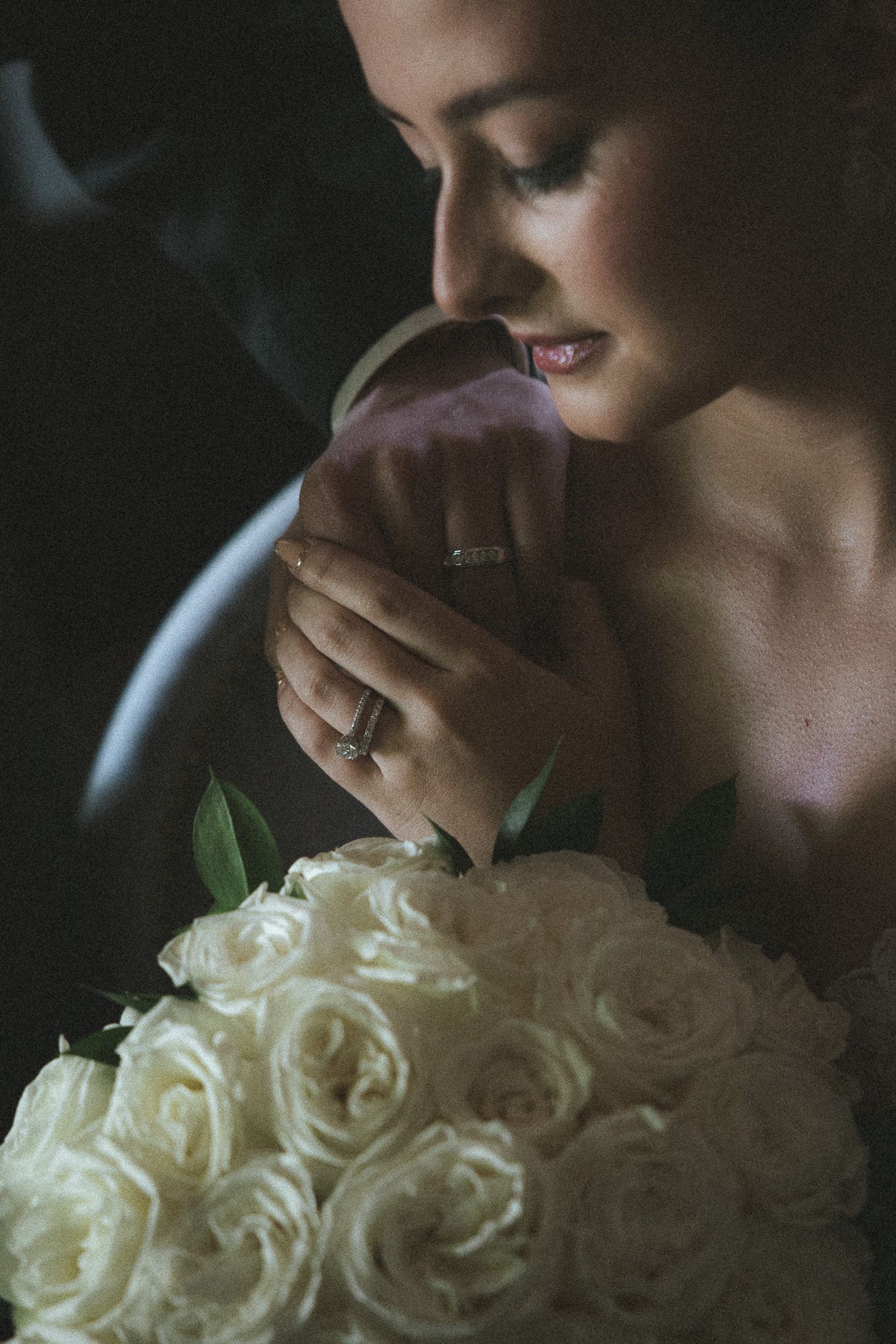 Close-up of bride smelling white rose bouquet while wearing diamond engagement ring in moody lighting.