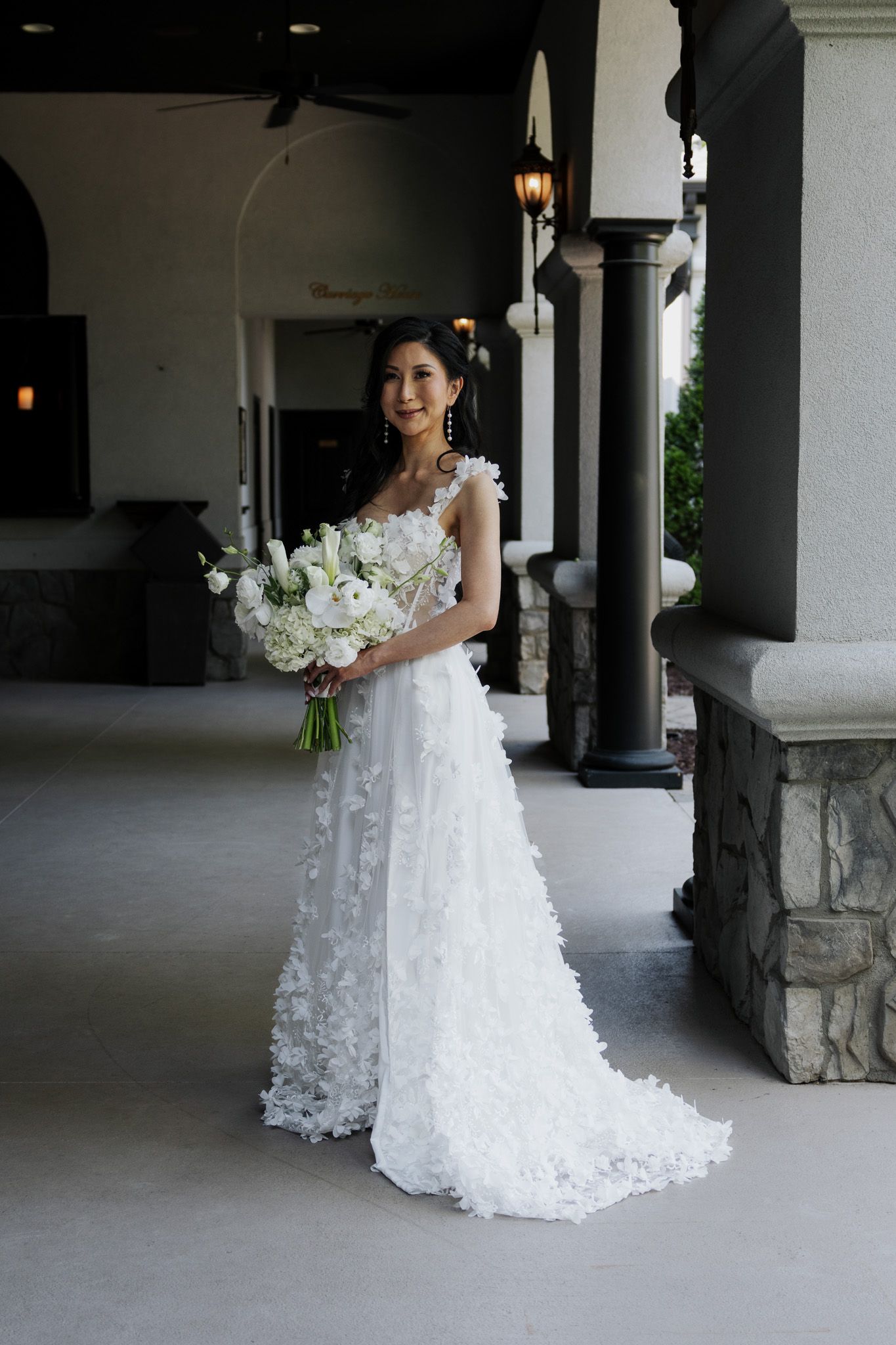 Smiling bride in 3D floral wedding dress holds white bouquet under covered venue entrance.