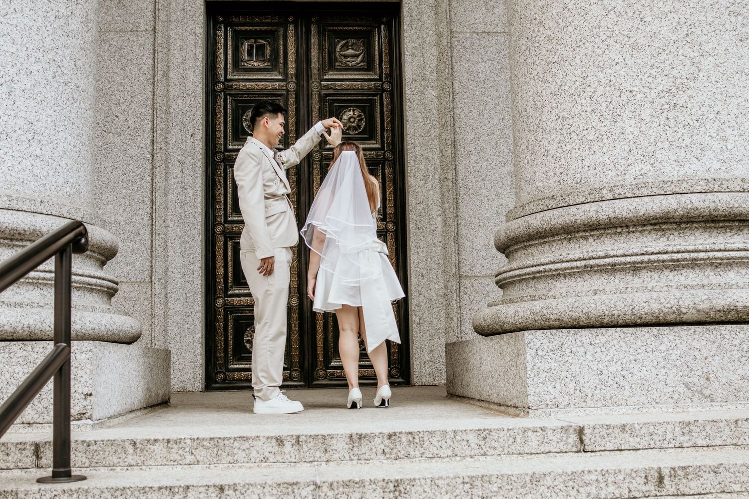 Bride and groom pose on steps of grand stone building as groom lifts bride's wedding veil.