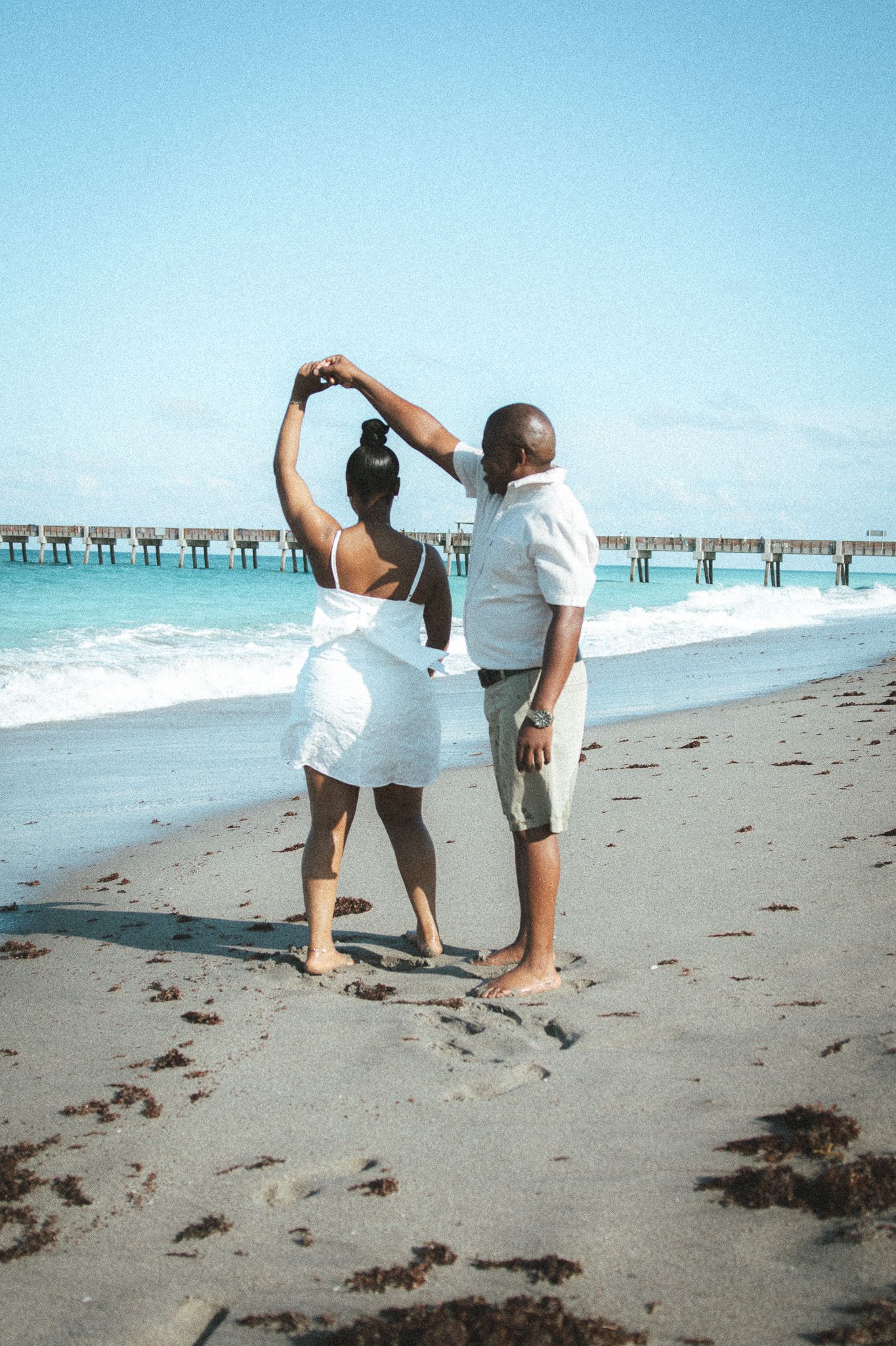 Couple in white outfits dance on a sandy beach, woman raises her arms gracefully with a turquoise ocean and pier behind.