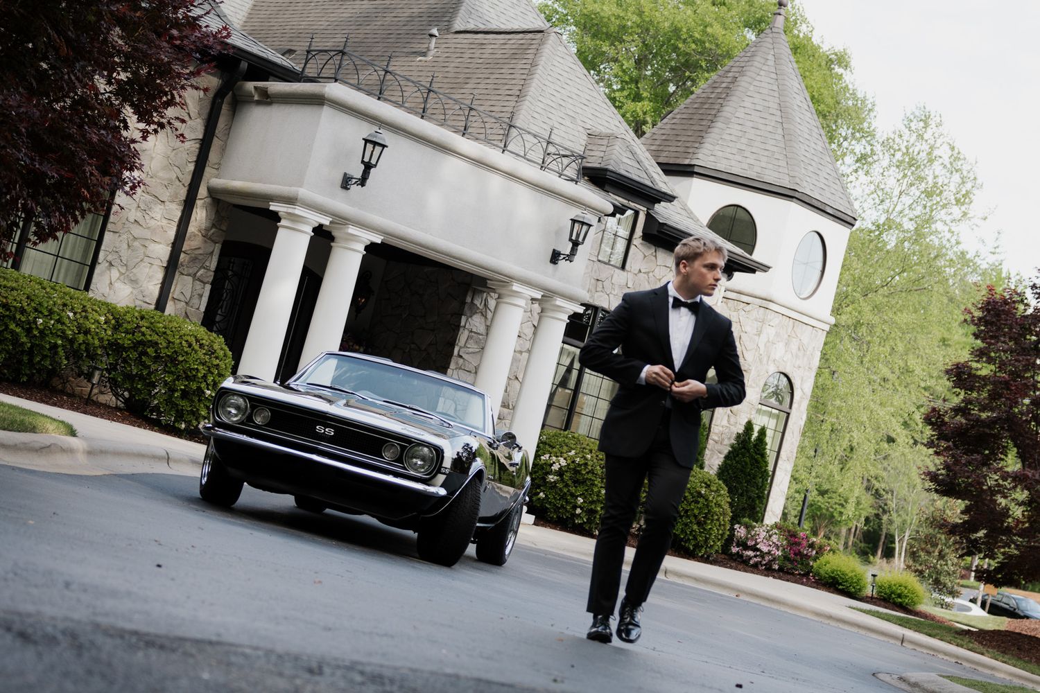 Groom in black tuxedo walks confidently down driveway with classic black muscle car and stone mansion behind him.