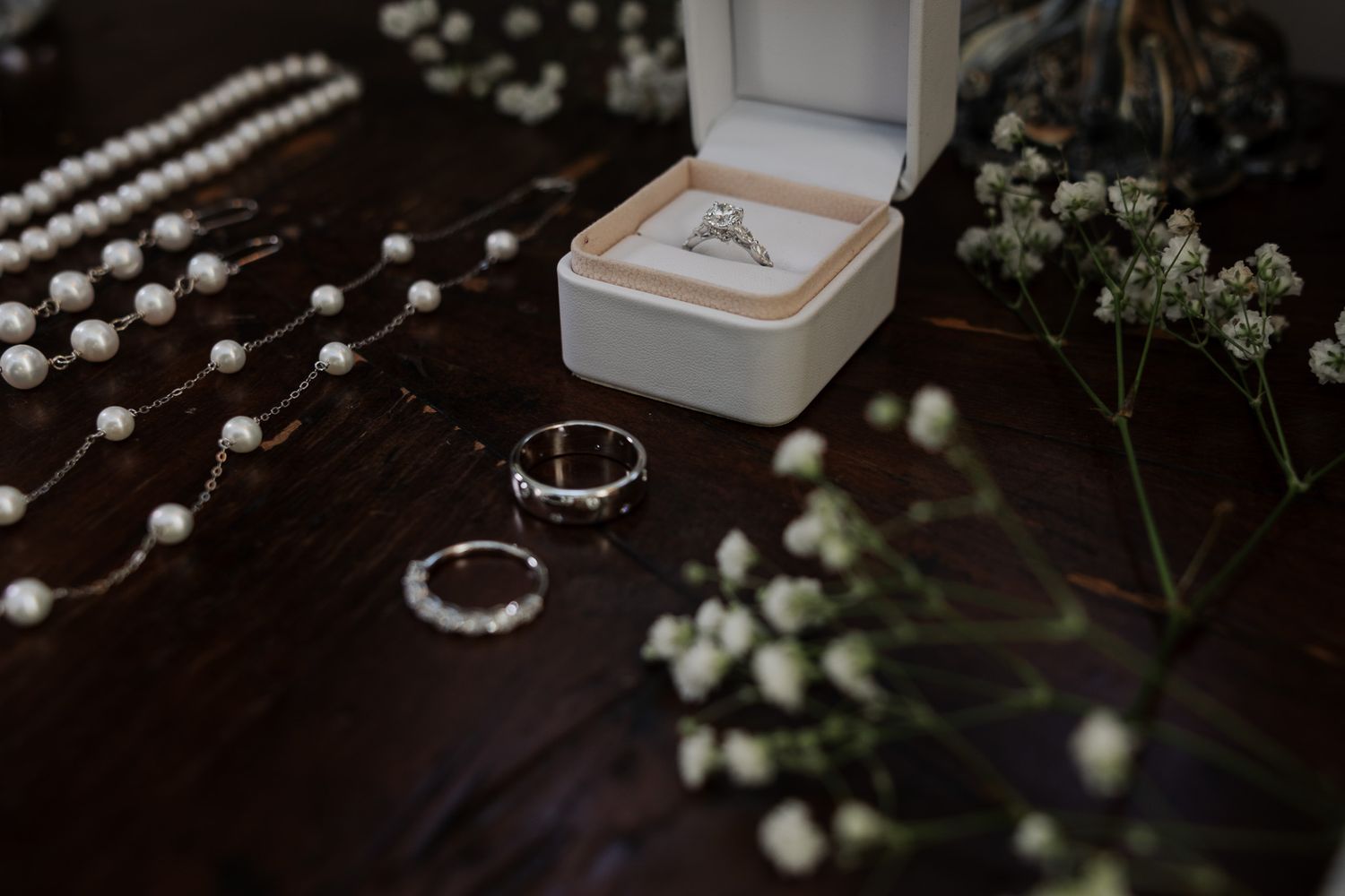 Wedding jewelry flat lay with diamond engagement ring in box, pearl necklace, and baby's breath flowers.