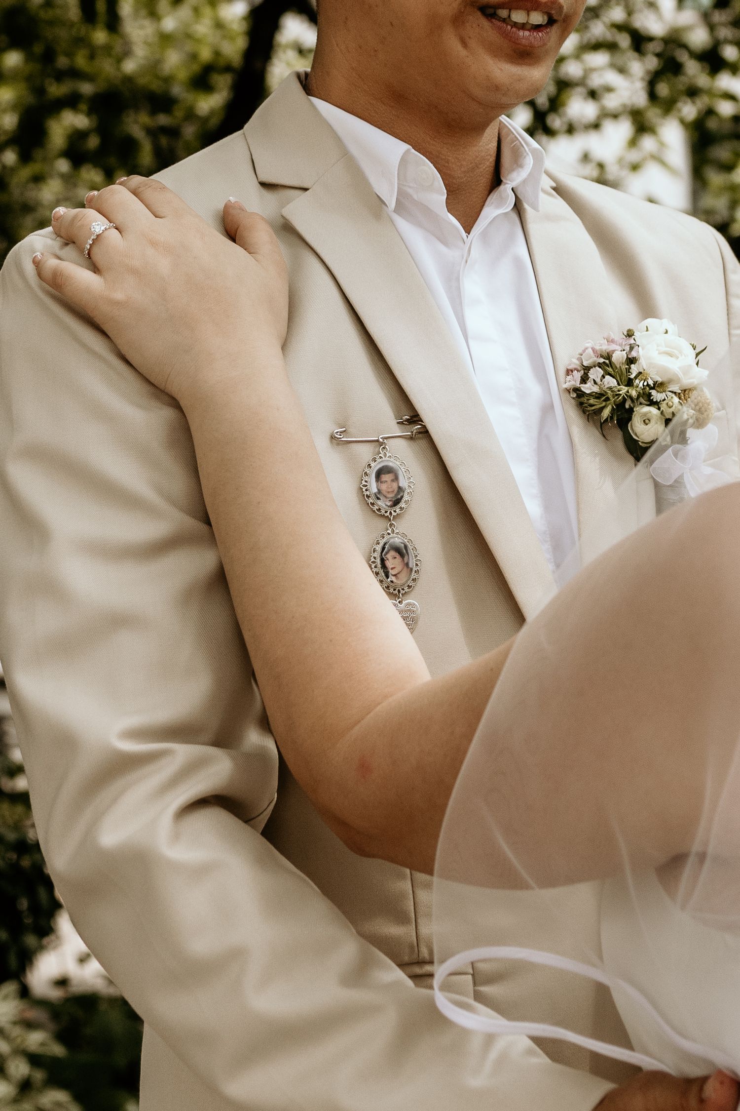 Bride embraces groom wearing beige suit with memorial photo pins and white boutonniere.