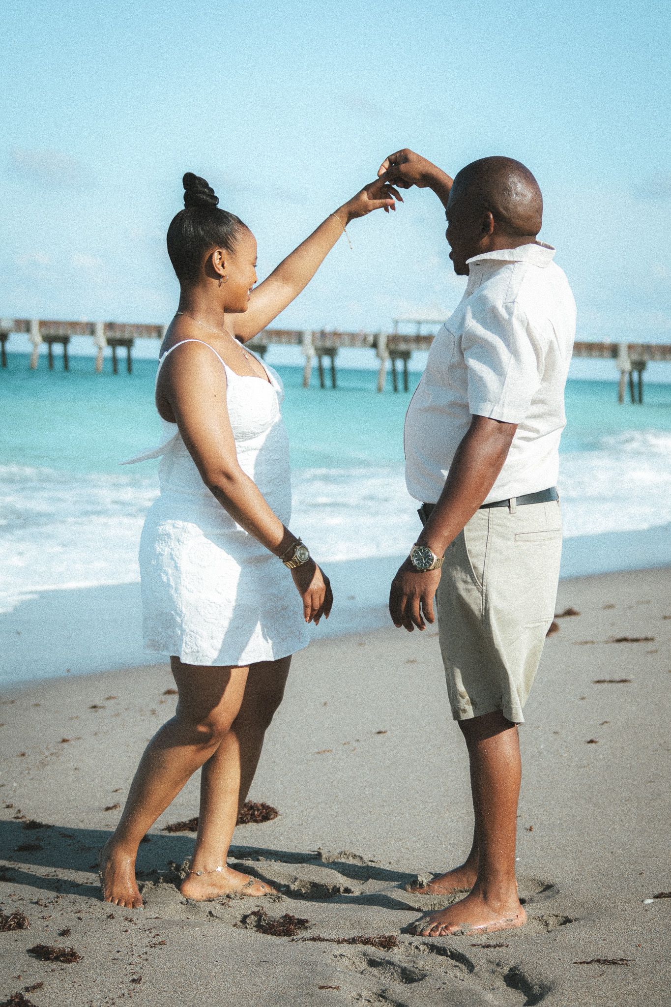 Woman gently touches her partners face as they dance together barefoot on a sunny beach with ocean waves behind them.