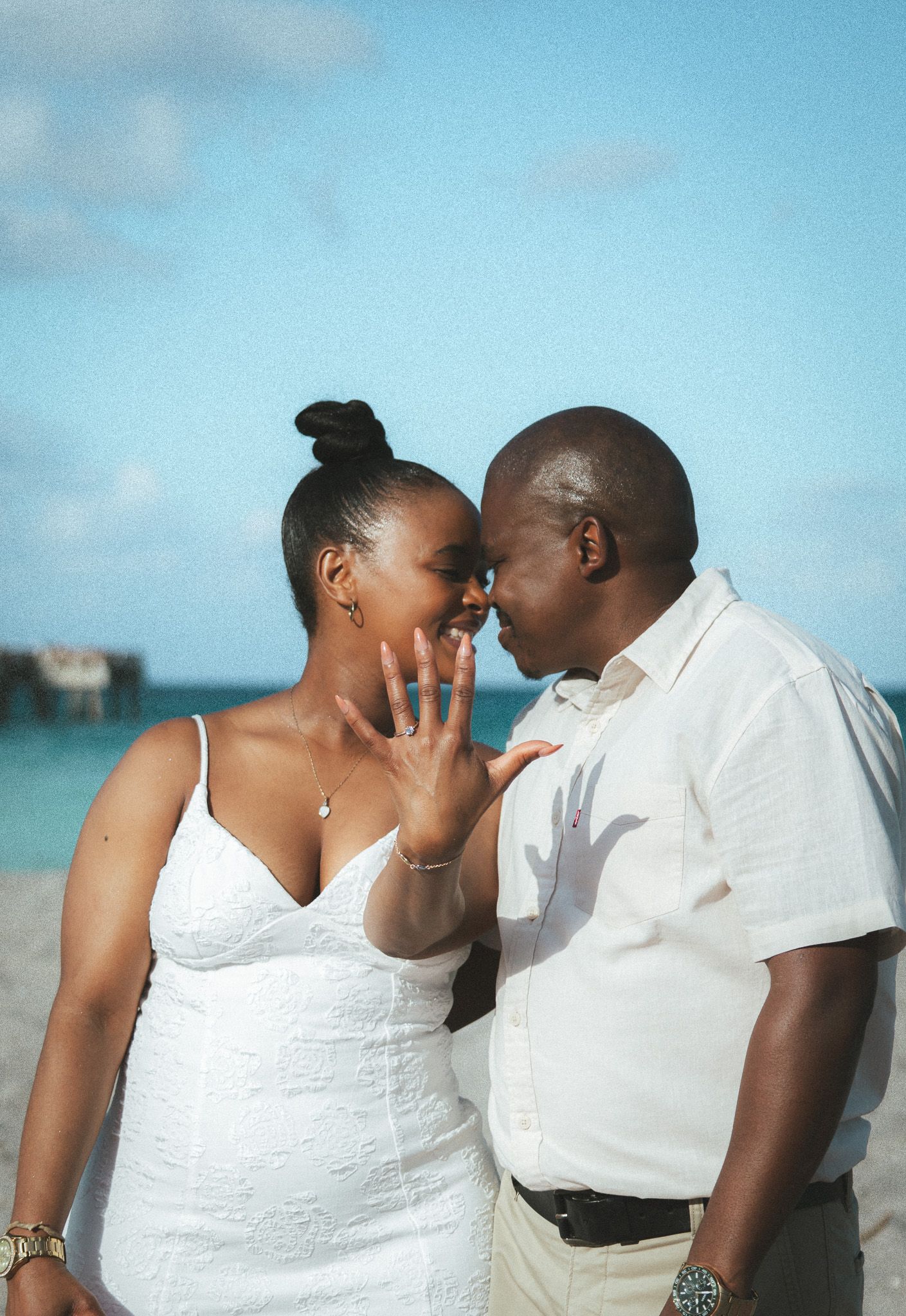 An engaged couple touching foreheads on the beach, with the woman proudly displaying her engagement ring to the camera.