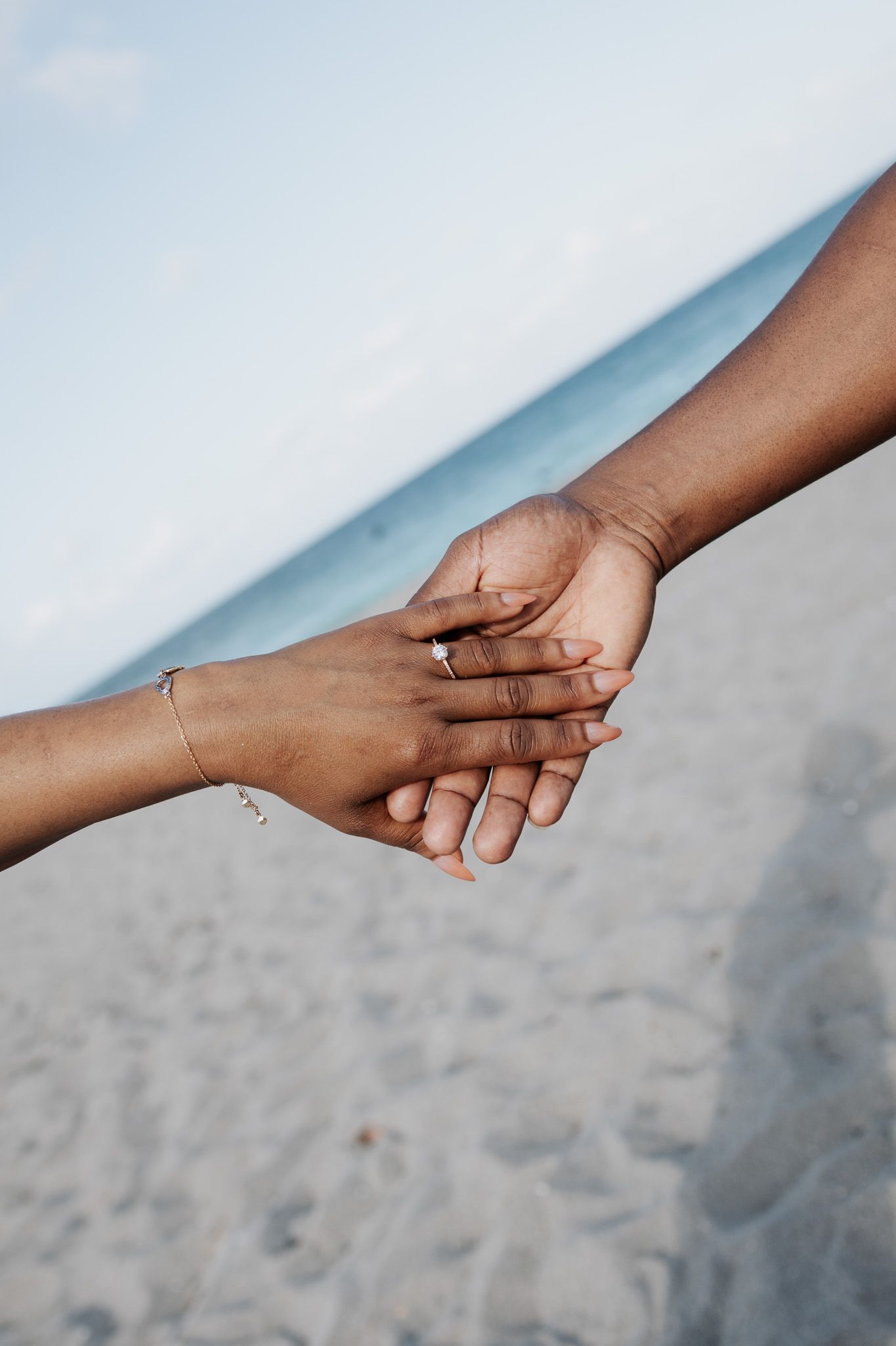 A close-up of an engaged couple holding hands on a sandy beach, showcasing a sparkling engagement ring on her finger.