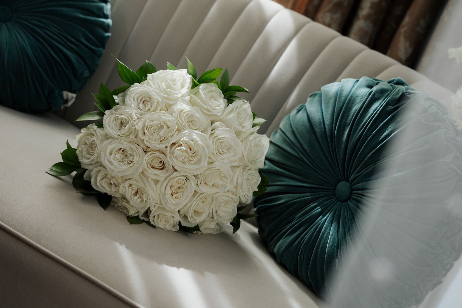White rose bridal bouquet resting on a silver chair beside teal velvet decorative pillows.