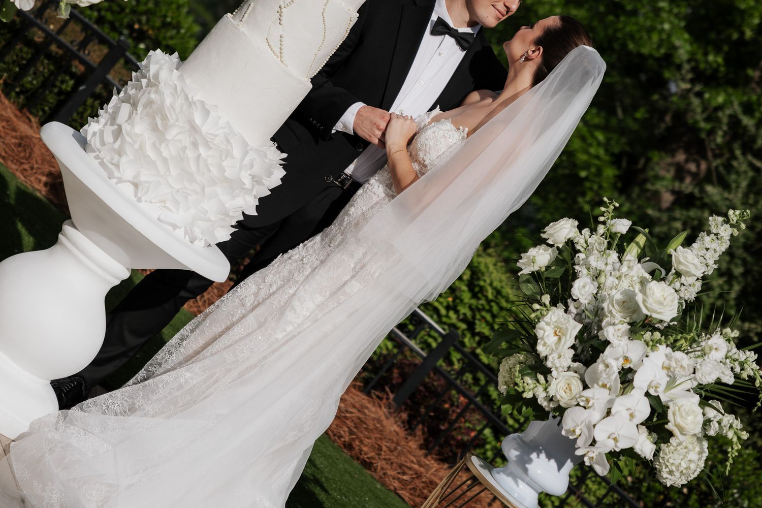 Bride in lace gown and groom in tuxedo embrace beside their tall white wedding cake surrounded by white flowers.