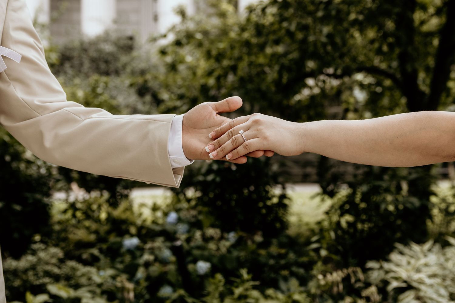 Close-up of bride and groom holding hands showing wedding rings with green garden background.