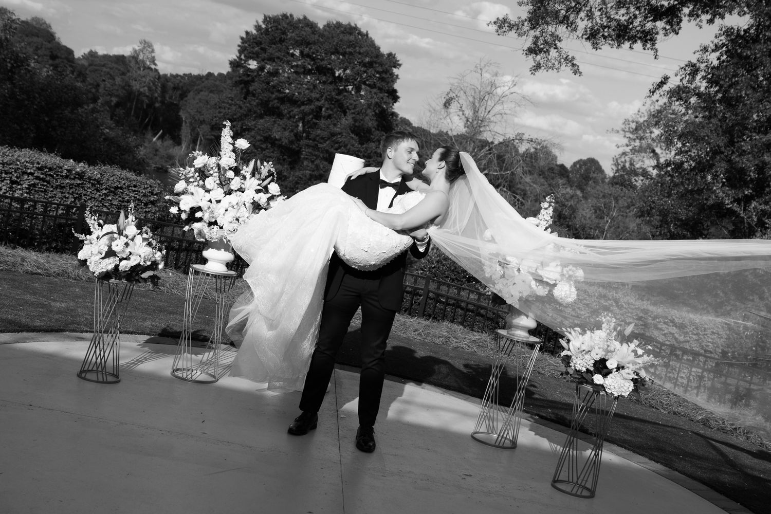 Black and white photo of groom carrying bride while kissing at outdoor wedding with floral decor.