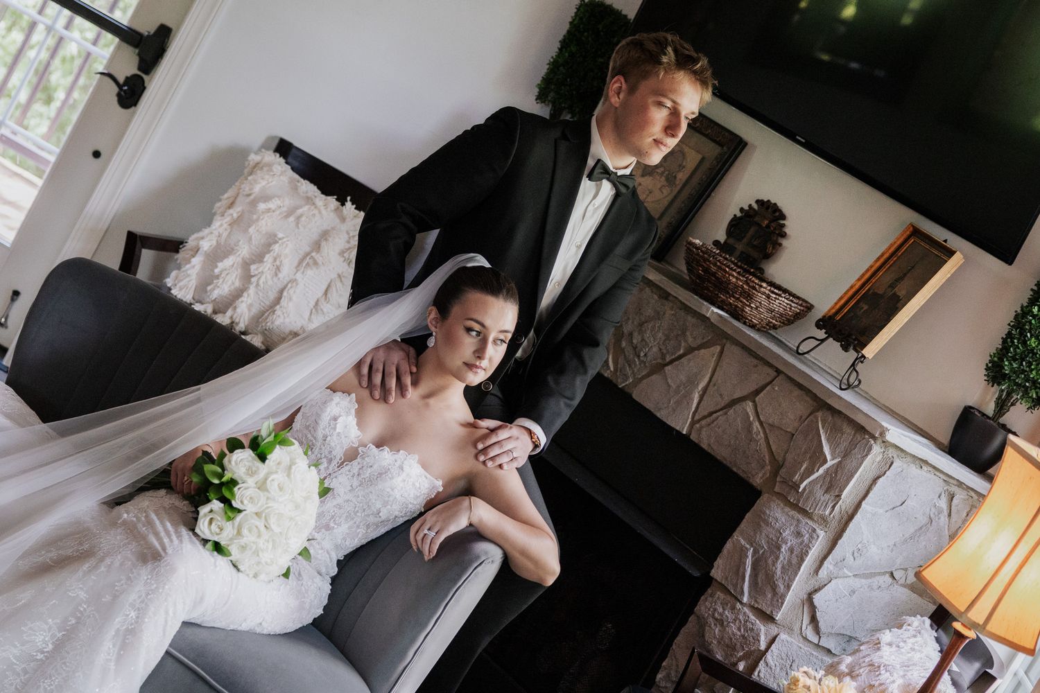 Bride and groom share an intimate moment on a gray sofa, with the groom in a black tuxedo standing behind her.