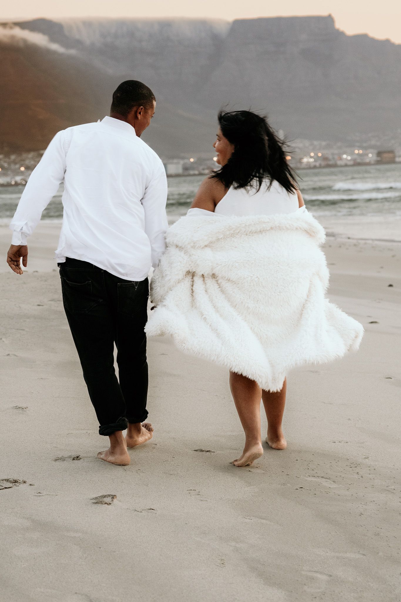 Couple walking barefoot on beach, man in white shirt and black pants, woman wrapped in white blanket, mountains in background.