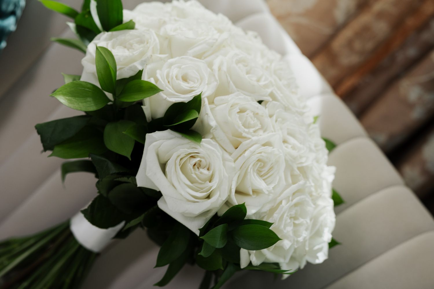 Elegant bridal bouquet of white roses with green leaves resting on a gray chair.